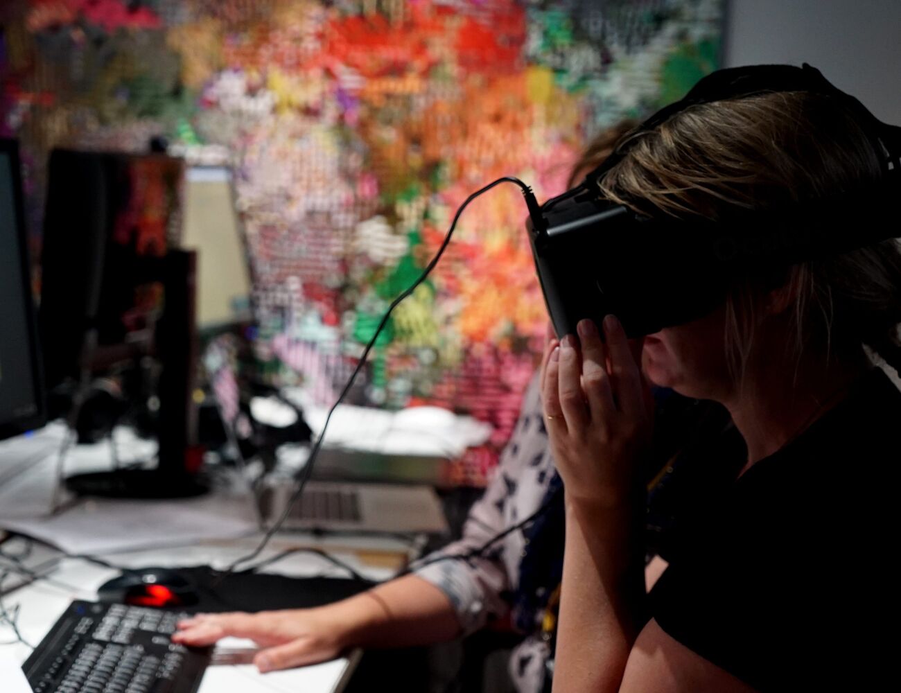 A person uses VR headset while another assists on the background