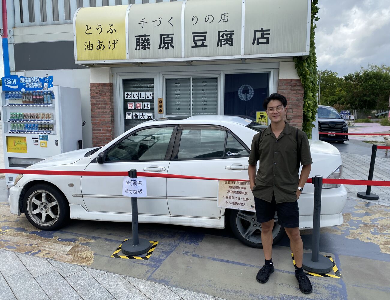 Songwen He seisoo valkoisen auton edessä Hongkongin kadulla. Taustalla kylttejä ja mainostekstejä kantoninkiinaksi.