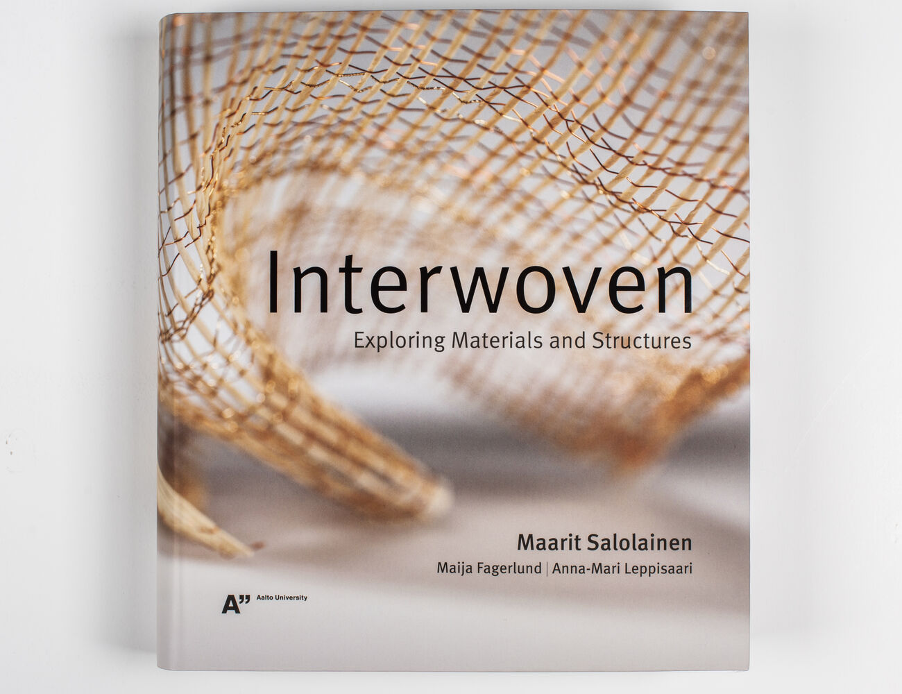 Interwoven-kirjan harmaansävyinen kansi, jossa on kuparilankainen kudos.
