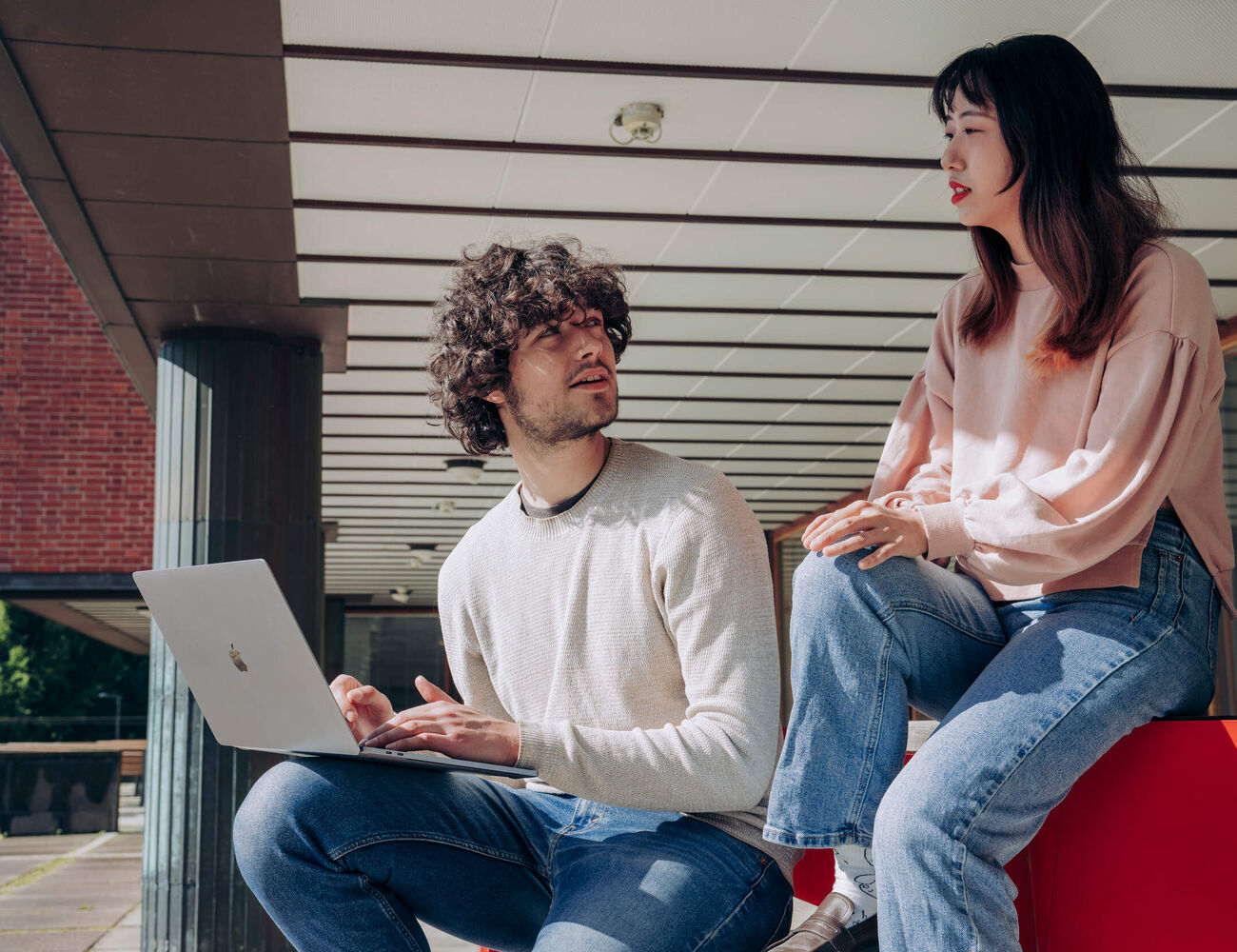 Two students sitting and discussing outside while one student is holding a laptop in his knee while listening to the other student.
