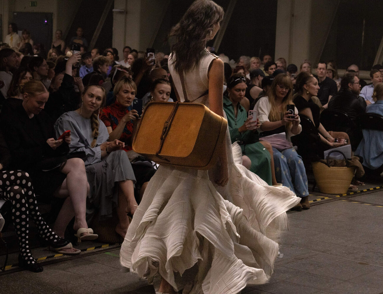 Model walks the catwalk in a light-coloured dress with a suitcase on her back