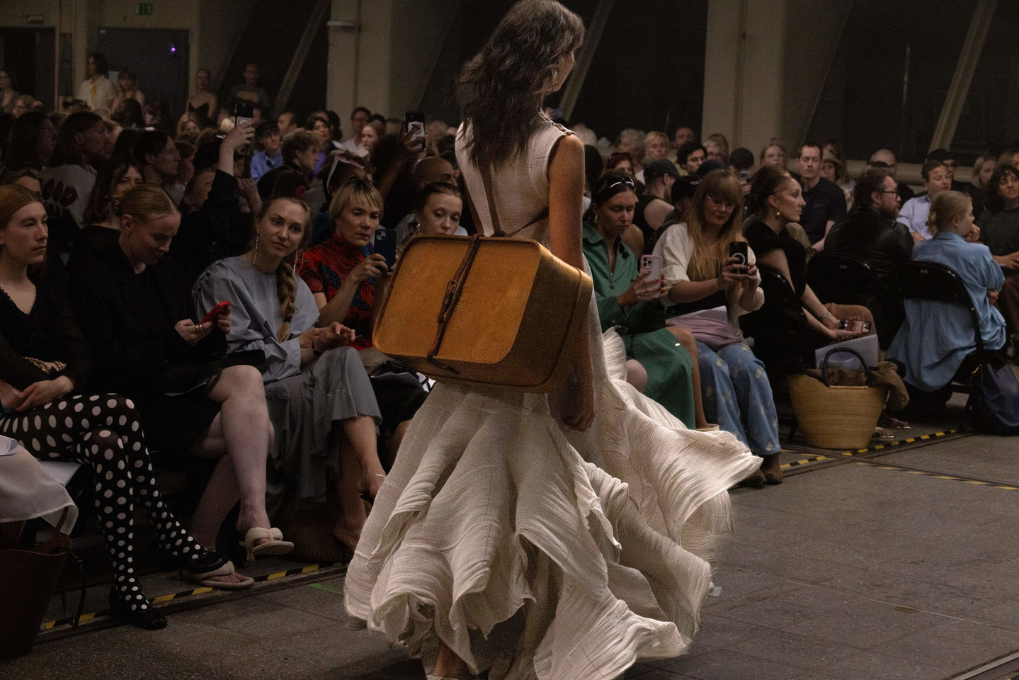 Model walks the catwalk in a light-coloured dress with a suitcase on her back