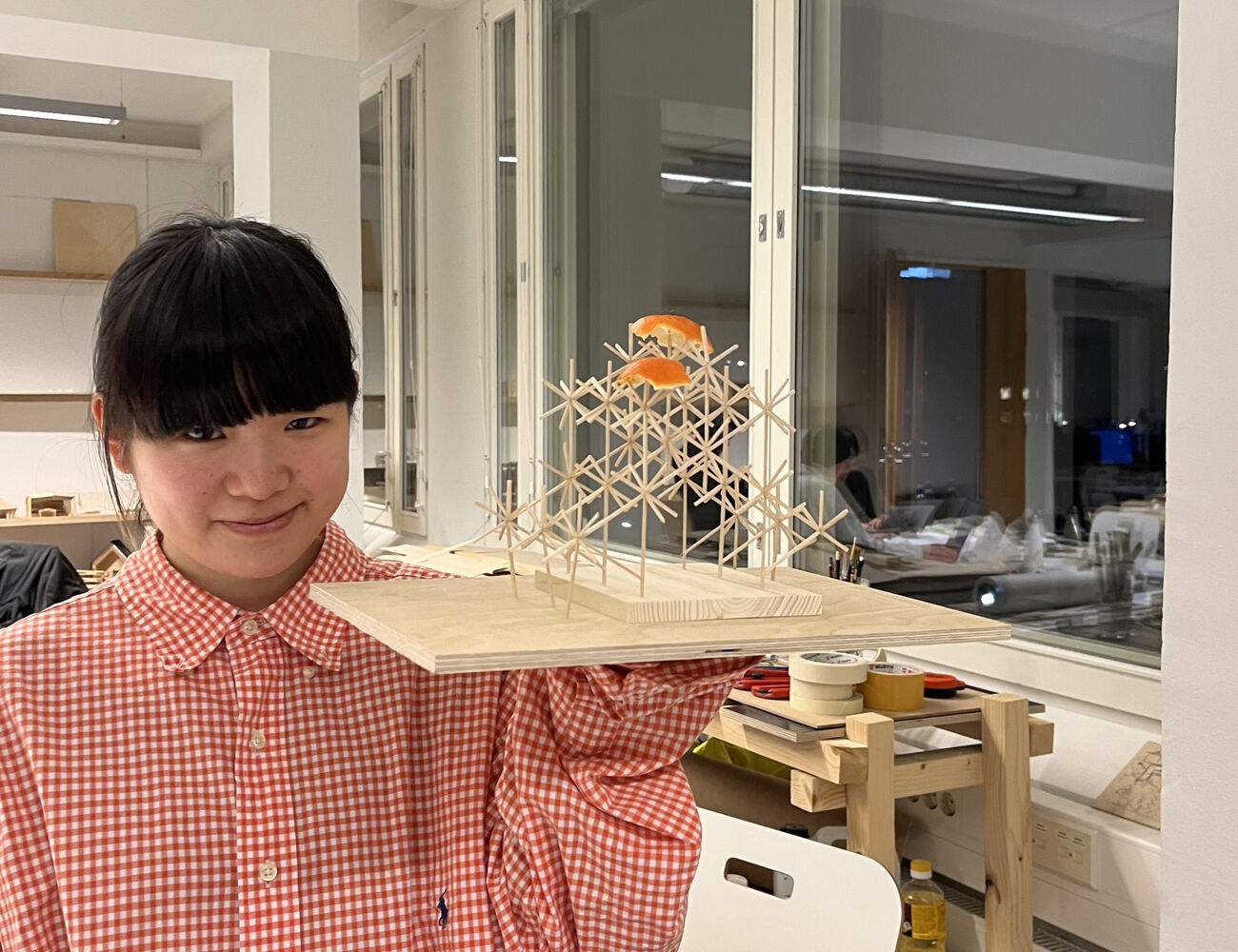 Student Meina holding up a wooden scale model of her design proposal, with two orange peels figuring as improvised roof