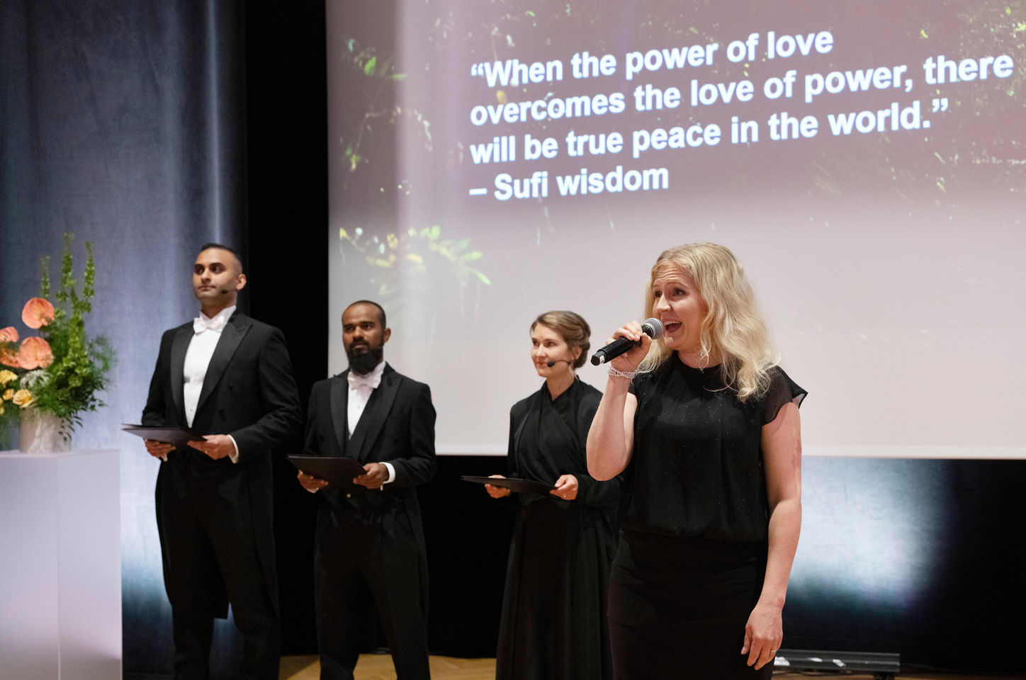 A women singing on a microphone, three people standing behind her in festive clothes