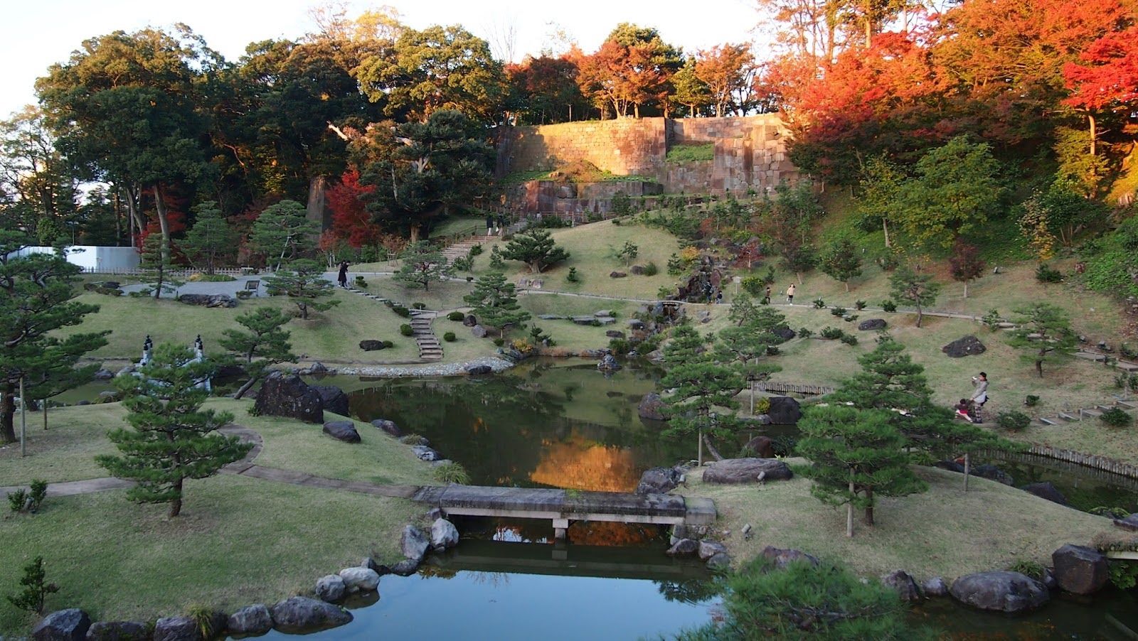 Puutarha, jossa on lampi, kivipolkuja ja syksyn lehtiä puissa. Ihmiset kävelevät ja nauttivat maisemasta.