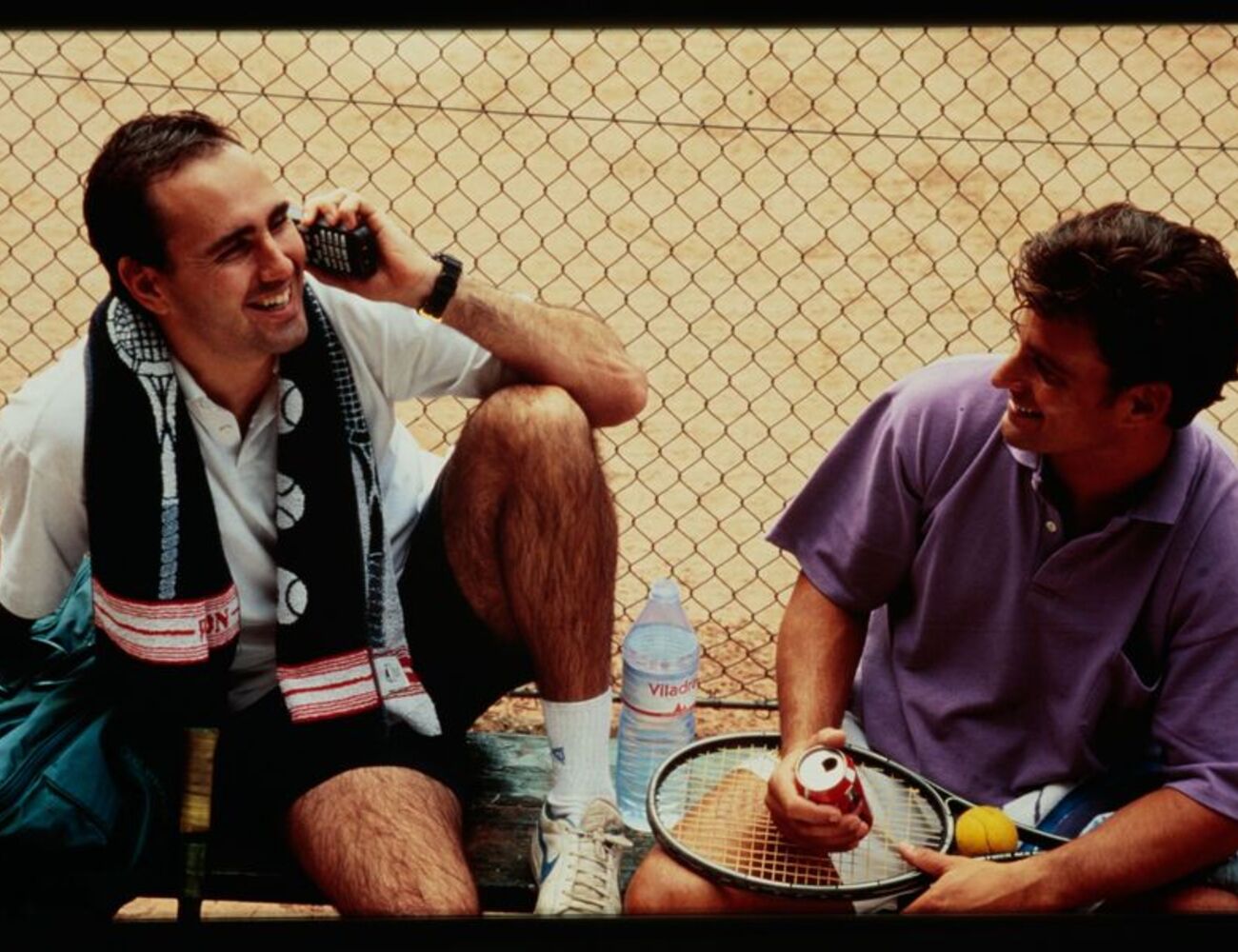 Two men sitting by a tennis court, one on the phone, the other holding a racket, with a water bottle and towel.