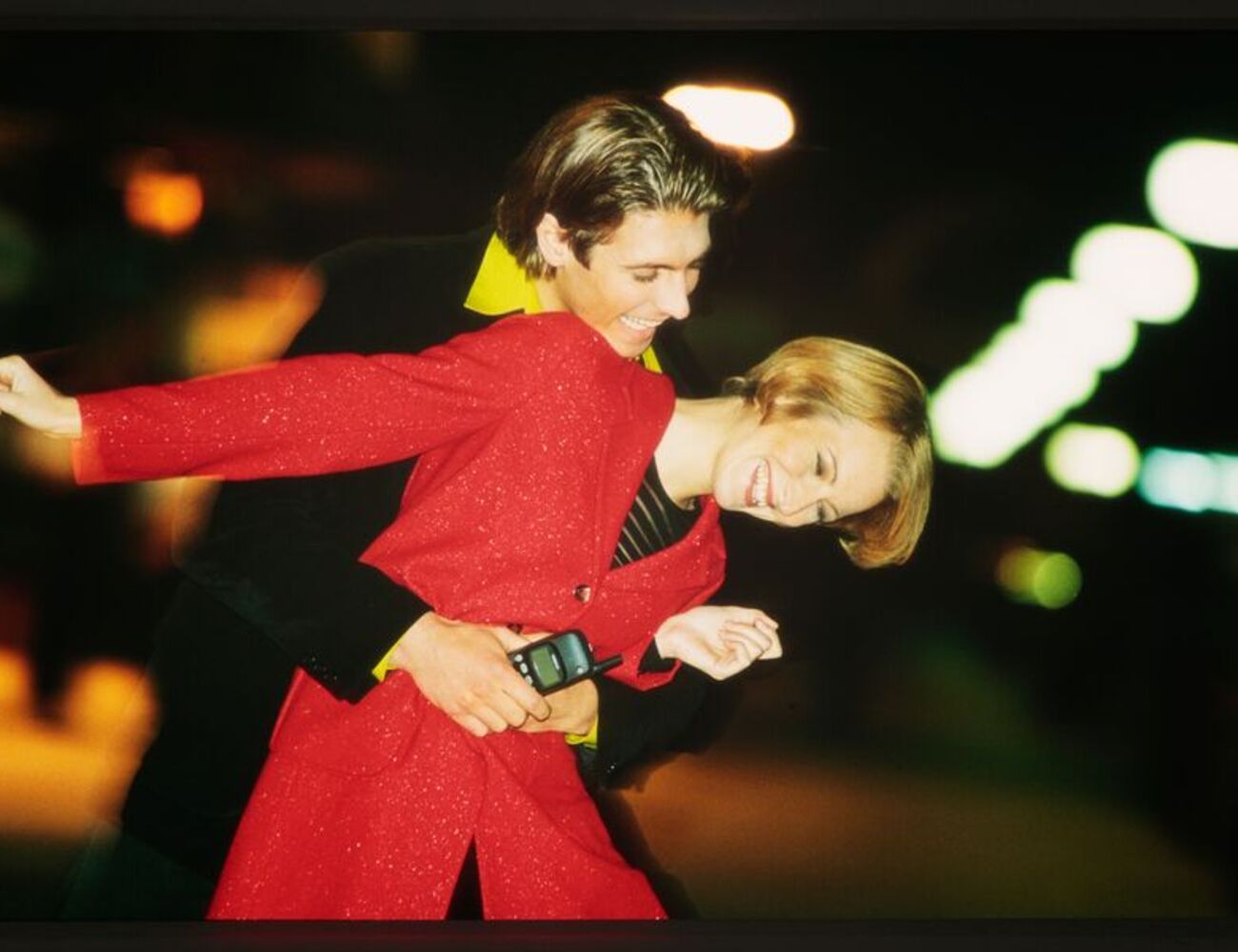 Two people, one in a red outfit, holding a phone, are posing closely together on a brightly lit street at night.