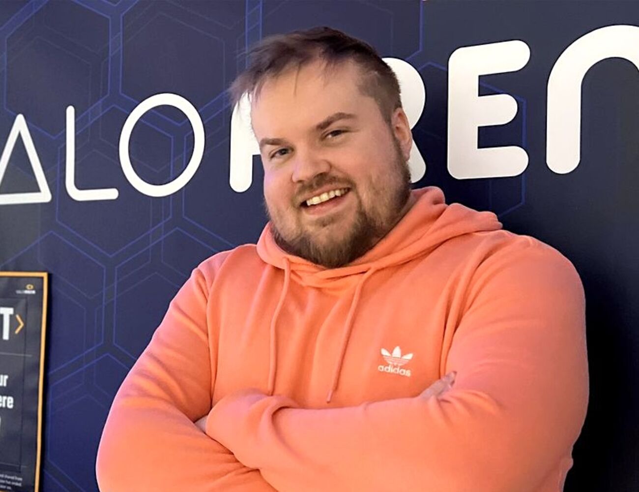 Oranssiin huppariin pukeutunut henkilö seisoo käsivarret ristissä seinän edessä, jossa on geometrisia kuvioita ja tekstiä.