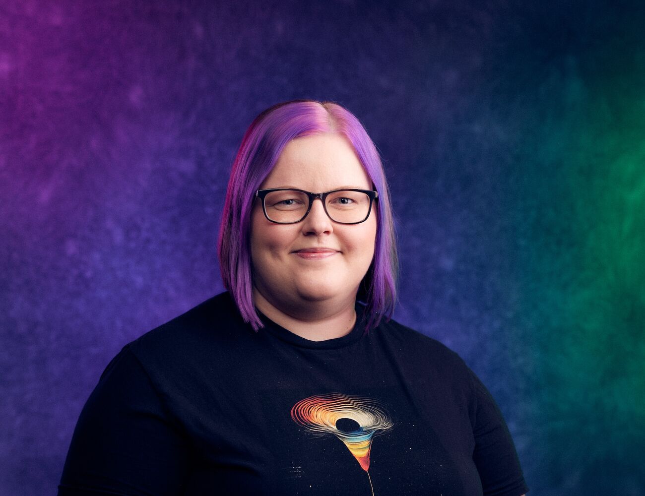 Henkilö, jolla on olkapäille ulottuvat hiukset ja musta paita, jossa on värikäs abstrakti kuvio. Tausta on liukuvärjätty violetti, sininen ja vihreä.