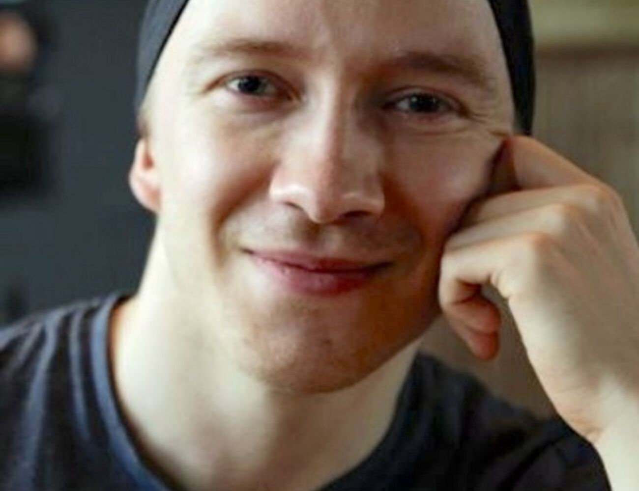 Henkilö, jolla on tumma paita ja hattu, istuu sisätiloissa ja nojaa kämmenselkäänsä.