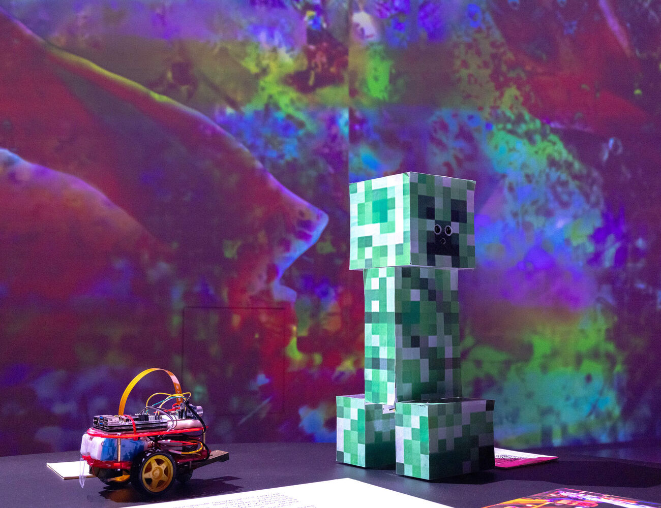 A Minecraft Creeper figure stands next to a small wheeled robot with a vivid, colourful background.