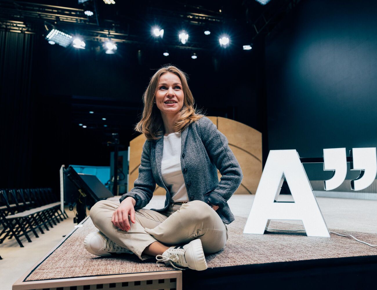Person sitting cross-legged on a stage next to a large 'A' and quotation mark sign, under stage lighting.