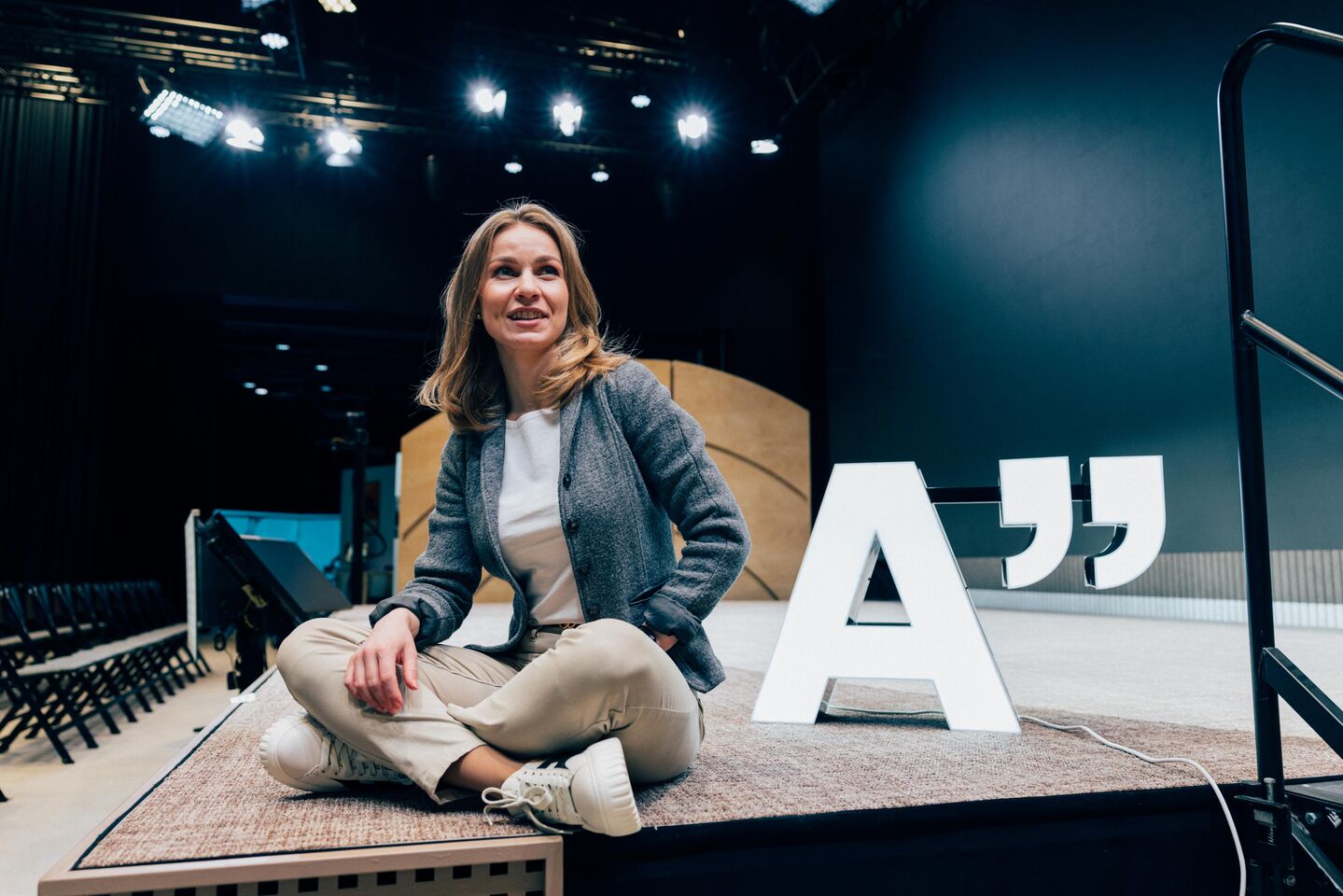 Henkilö istuu ristissä jaloin lavalla suuren 'A' ja lainausmerkkikyltin vieressä, näyttämövalaistuksessa.