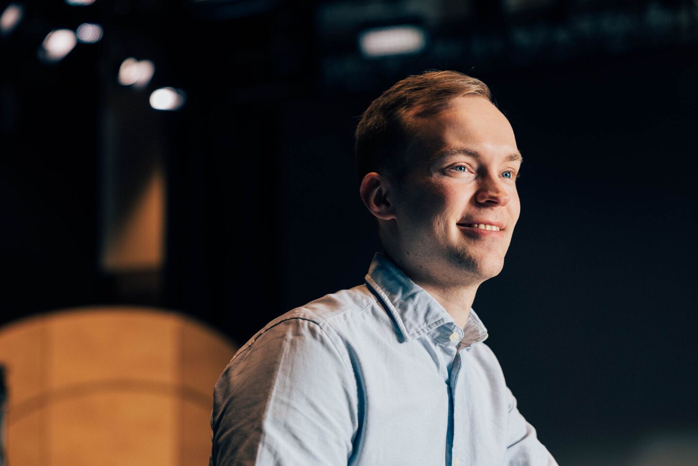 Henkilö vaaleansinisessä paidassa seisoo sisätiloissa tummalla taustalla, yläpuolella on valoja.