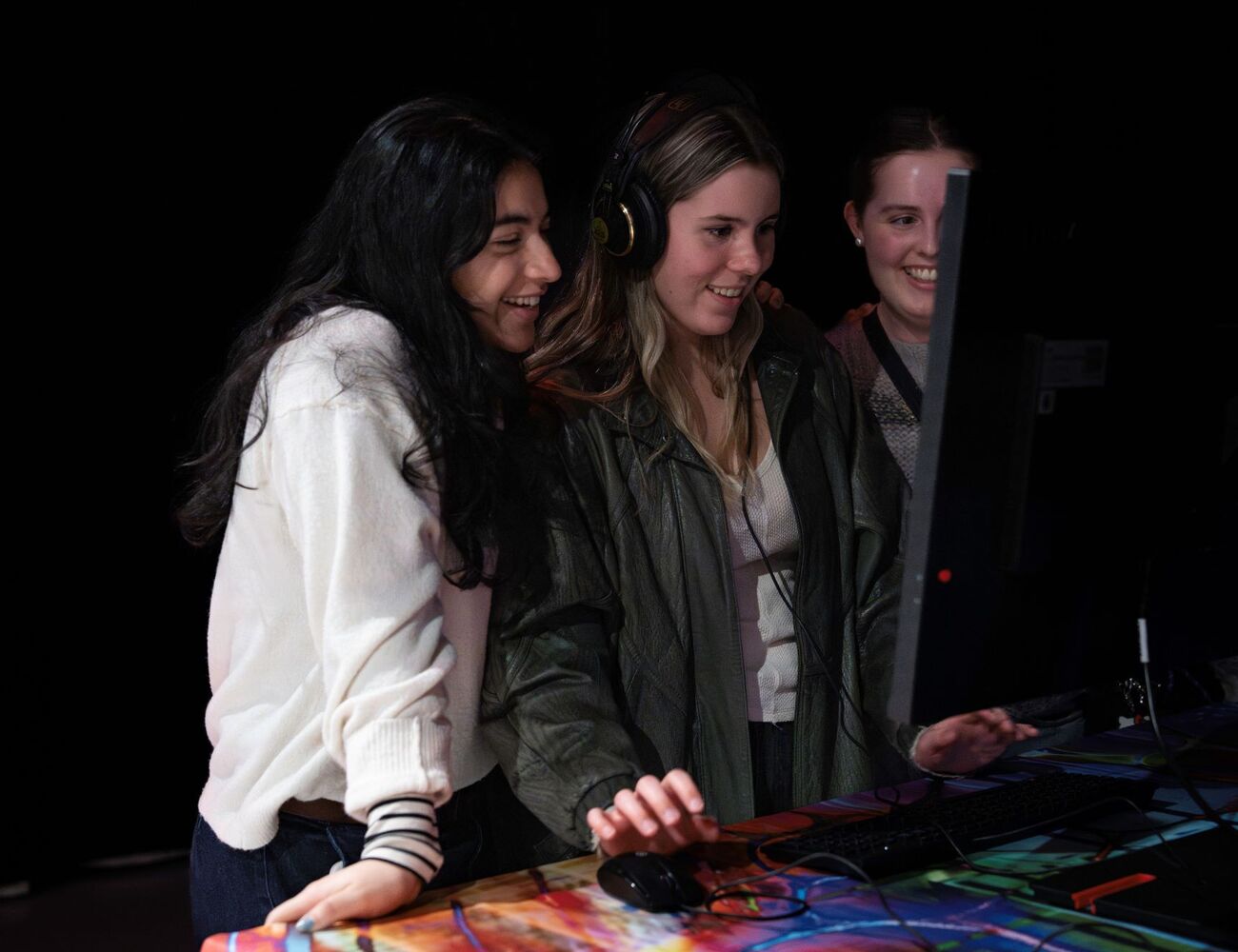 Three people are gathered around a computer screen, with one person wearing headphones and using a mouse.
