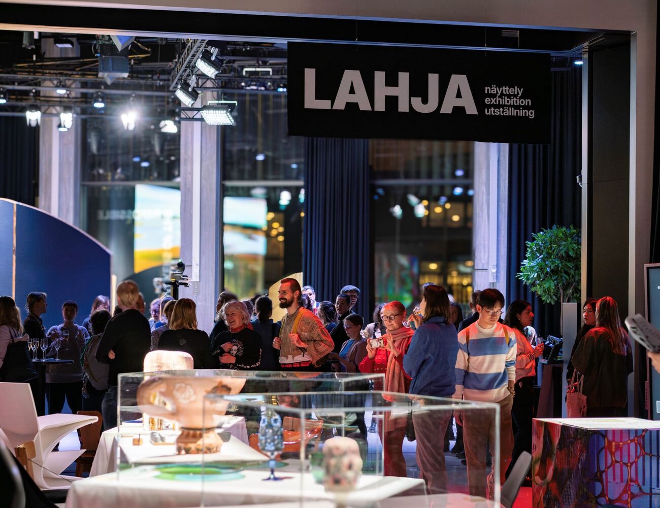 People attending a gift exhibition with various artefacts displayed. A plant and digital screen are in the background.