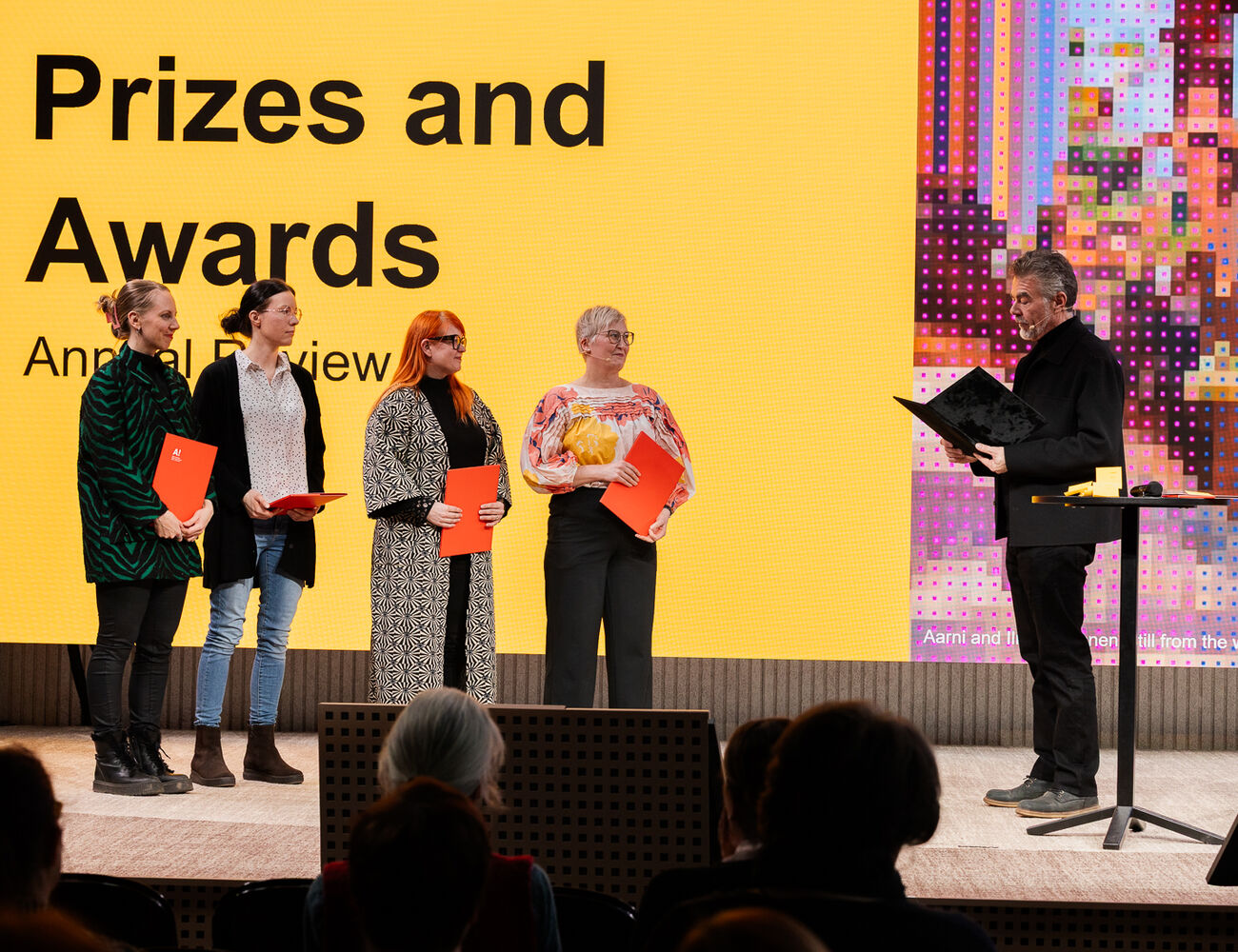 Four people on stage holding red certificates, with a man presenting an award. Background shows 'Prizes and Awards'.