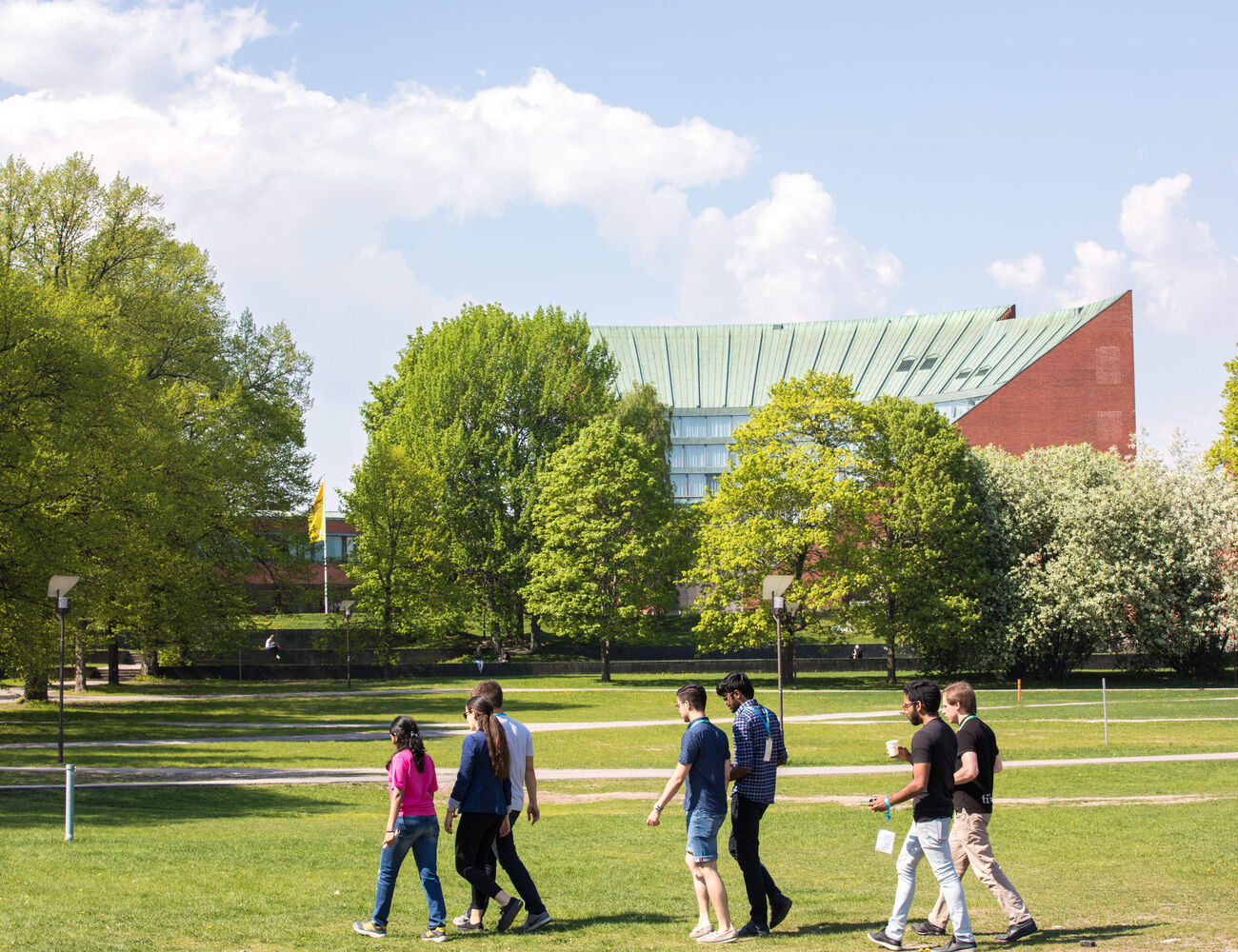 Group of people walking across a green park with trees. Aalto's buildings in the background.