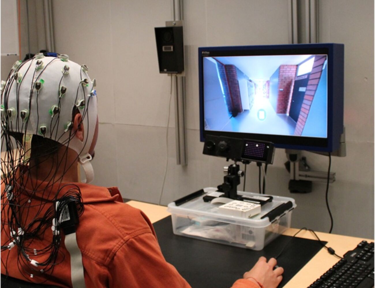 A person wearing an EEG cap sits at a computer, viewing a display with a virtual corridor.