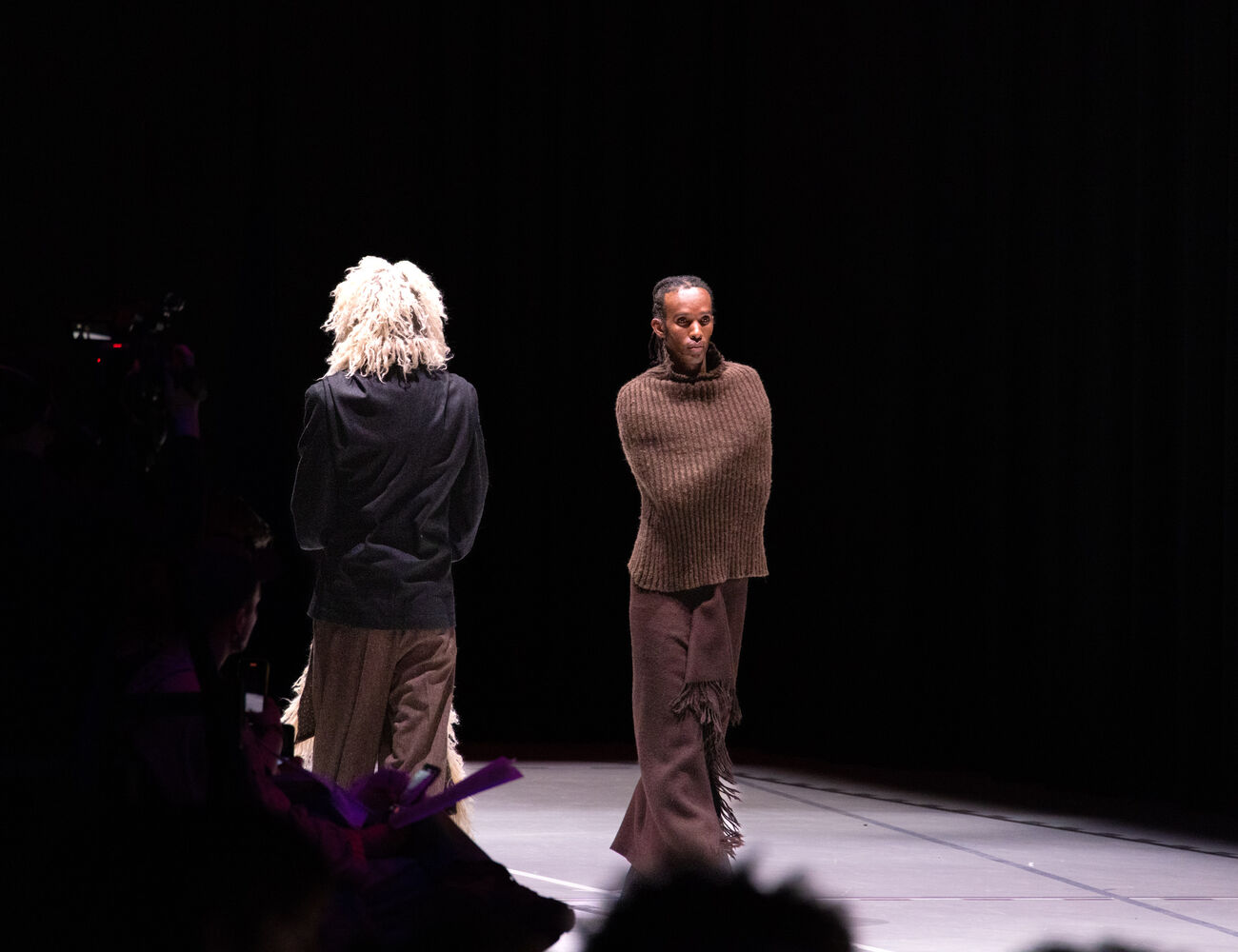 Two models on stage wearing unique brown and beige outfits under spotlight, with a dark background.