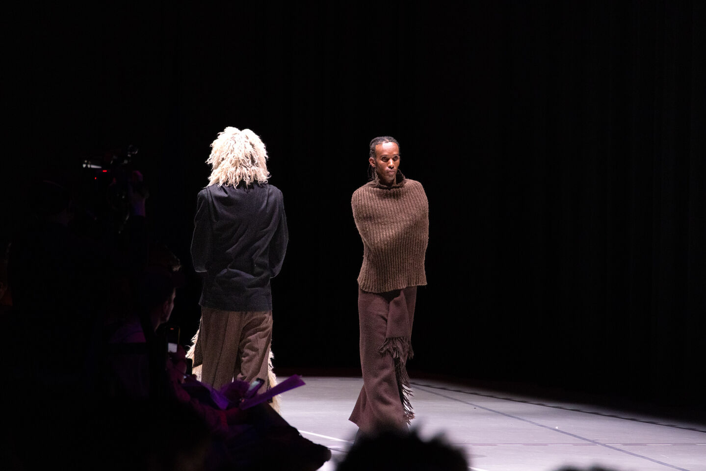 Kaksi mallia lavalla pukeutuneena erikoisiin ruskeisiin ja beigeihin asuihin, valokeilassa, tumma tausta.