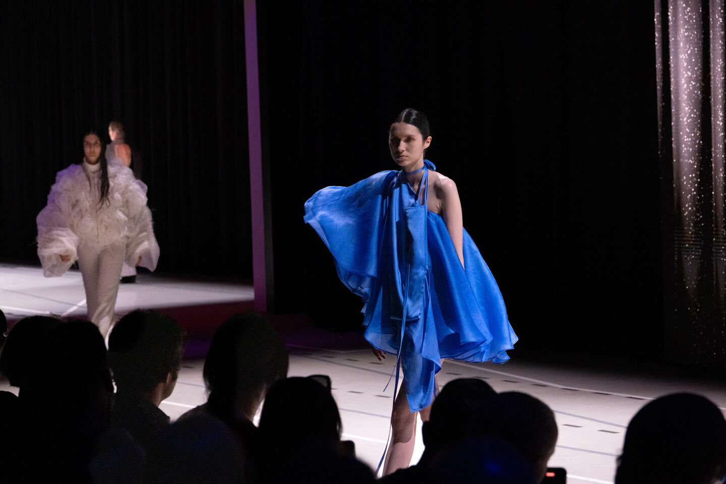 Kaksi mallia catwalkilla, toinen sinisessä ilmavassa mekossa, toinen valkoisessa teksturoidussa asussa, yleisö katsoo.