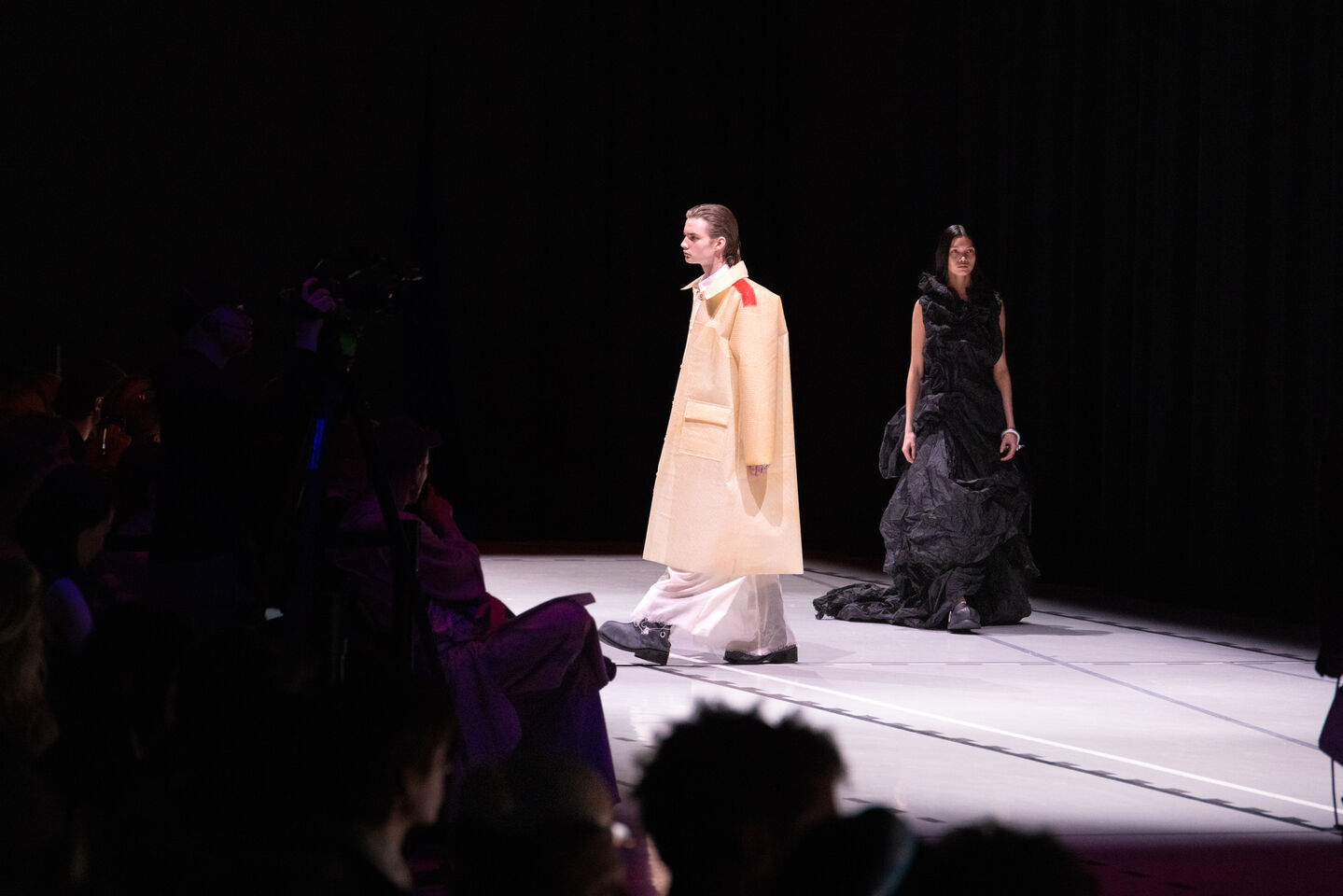 Kaksi mallia kävelee catwalkilla, toinen beige asu yllään ja toinen mustassa mekossa, yleisö katsoo.
