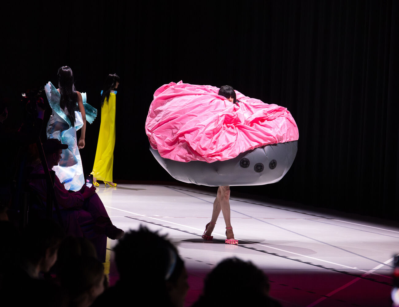 A model in a pink and grey, oversized, avant-garde outfit walks a runway. Other models and the audience are visible.