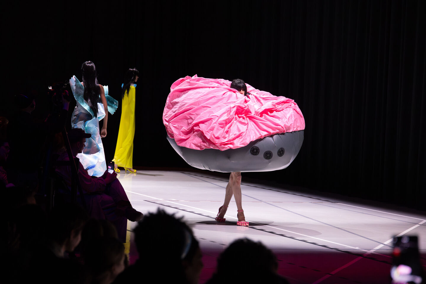 Malli pinkissä ja harmaassa, ylisuureessa avantgarde-asussa kävelee catwalkilla. Muita malleja ja yleisö näkyvissä.