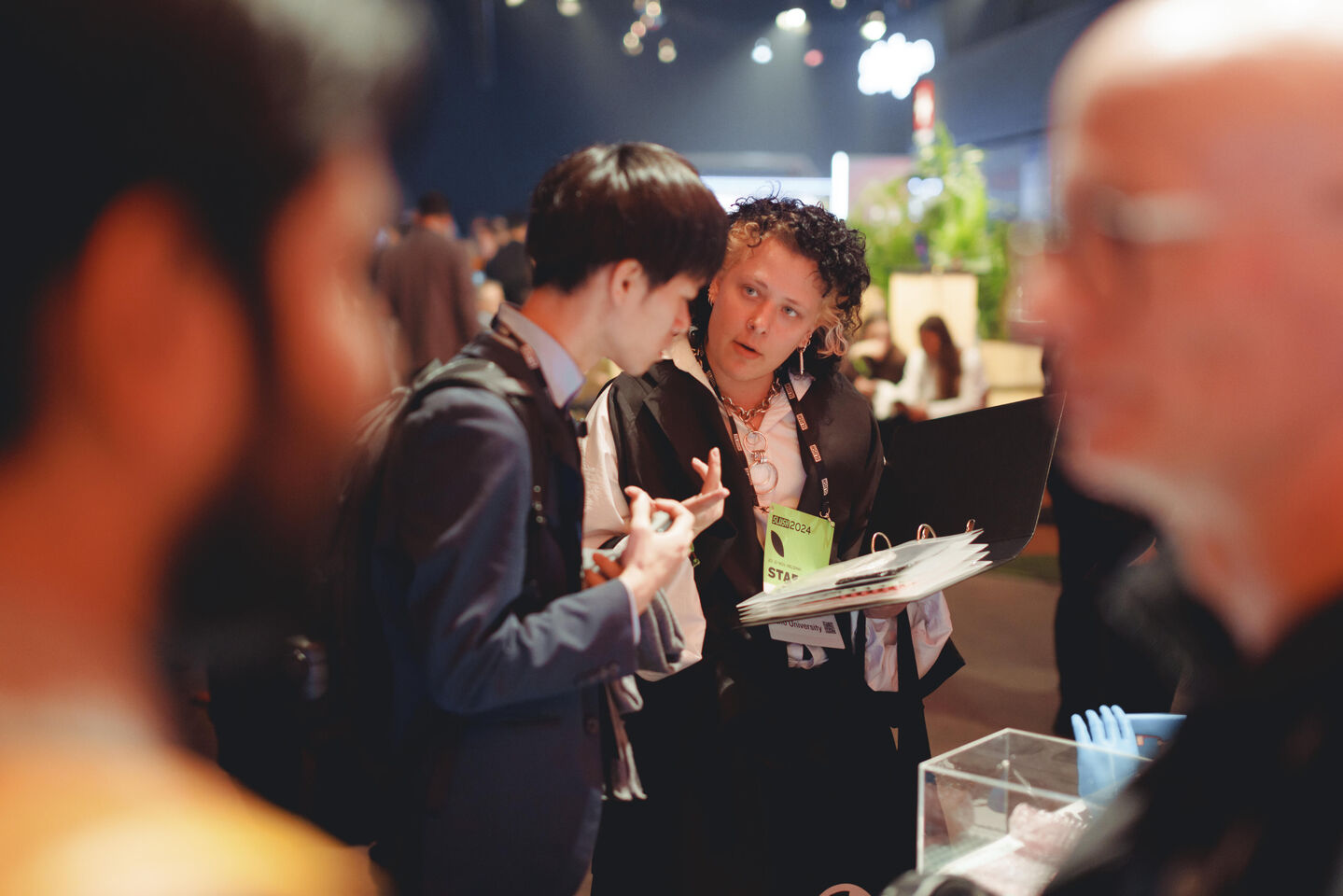 Two people in conversation at an indoor event. One holds a folder and the other gestures with their hand.