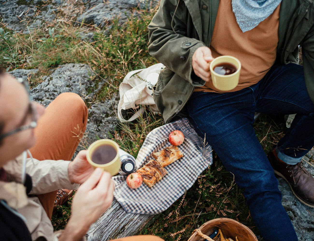 Two people picnicking outdoors, holding coffee mugs. On the cloth are pastries, an apple, and a camera.
