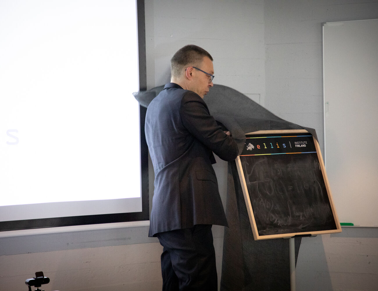 Man in a suit unveiling a blackboard next to a large whiteboard and projector screen.