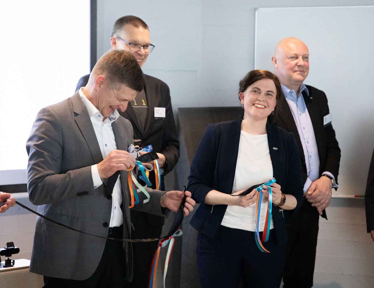 Four people in business attire hold colourful ribbons at an indoor event.