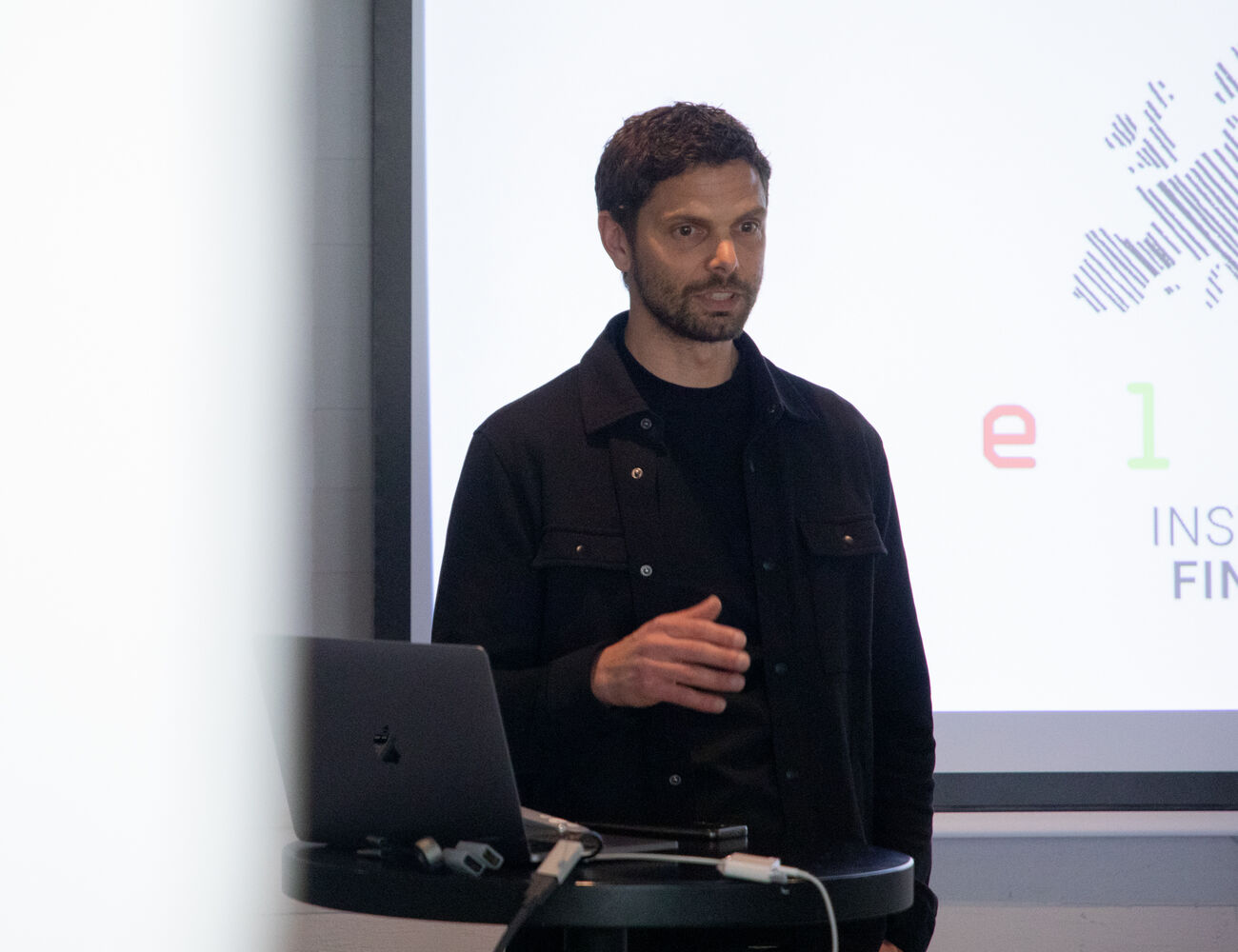 A person wearing a black outfit gives a presentation with a laptop on a table. A large screen shows text on the right.