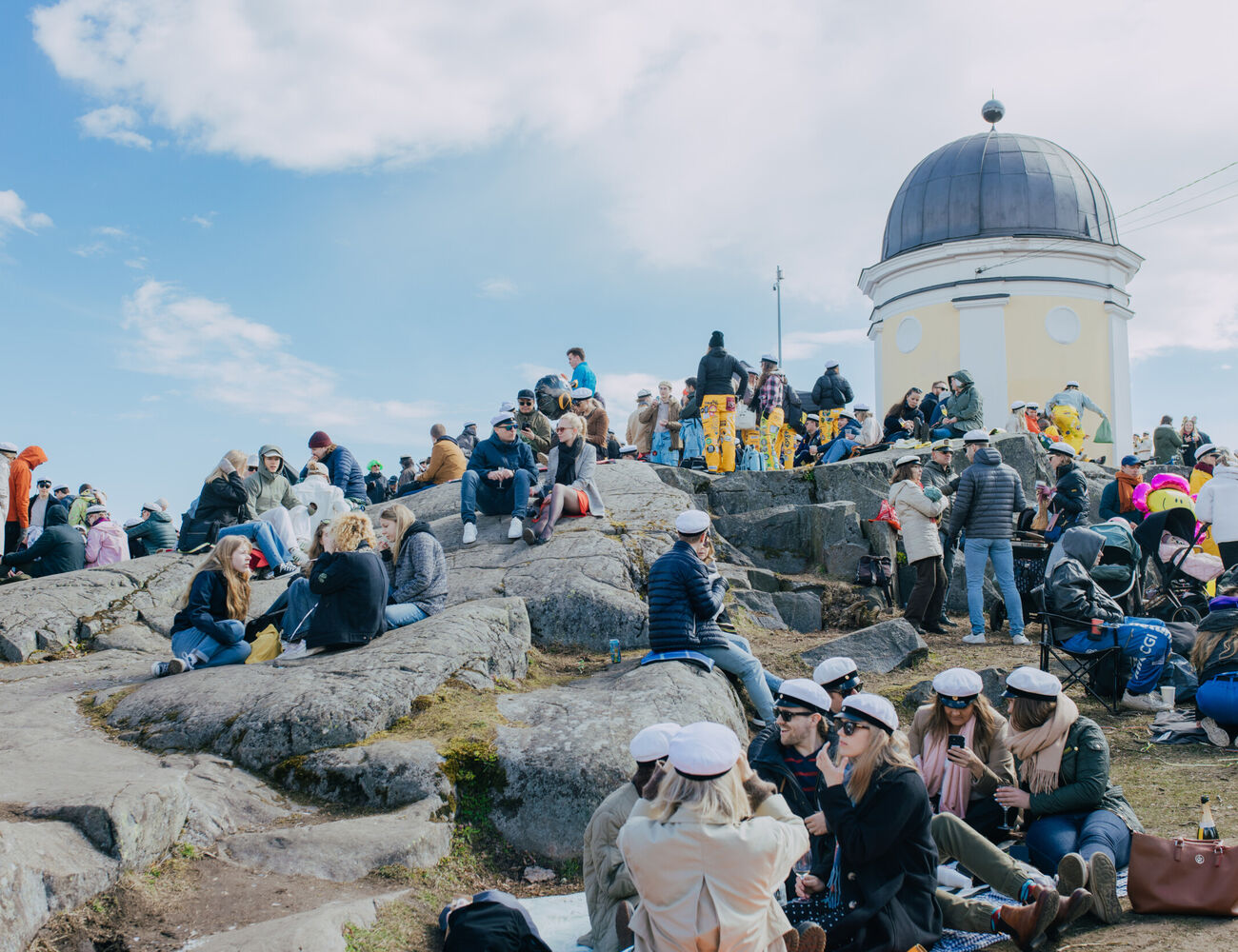 A large group of students celebrating outdoors on rocky terrain near a yellow observatory tower, many wearing white caps.