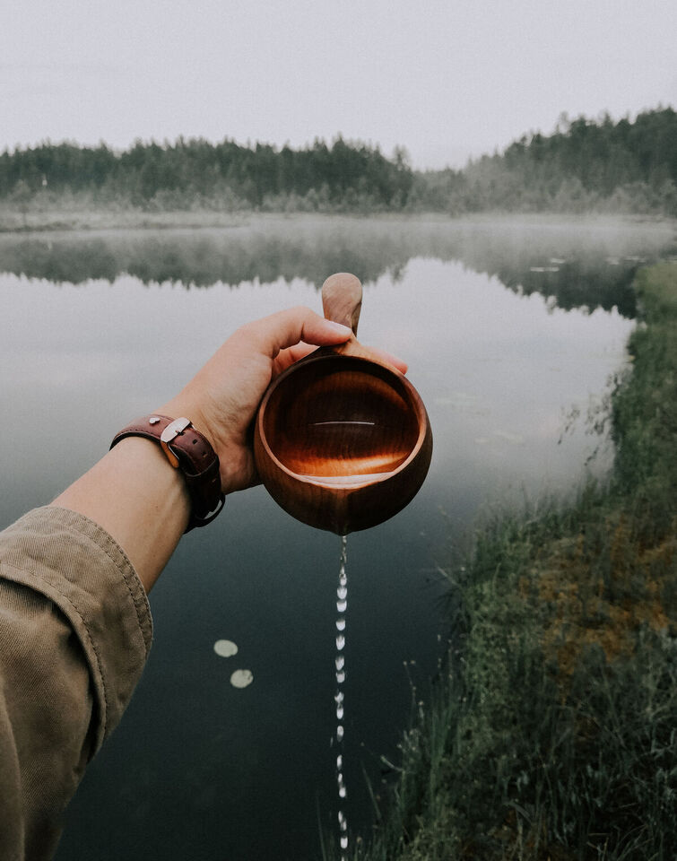 A hand holds a wooden cup, pouring water out into a lake, surrounded by misty forest and reeds.