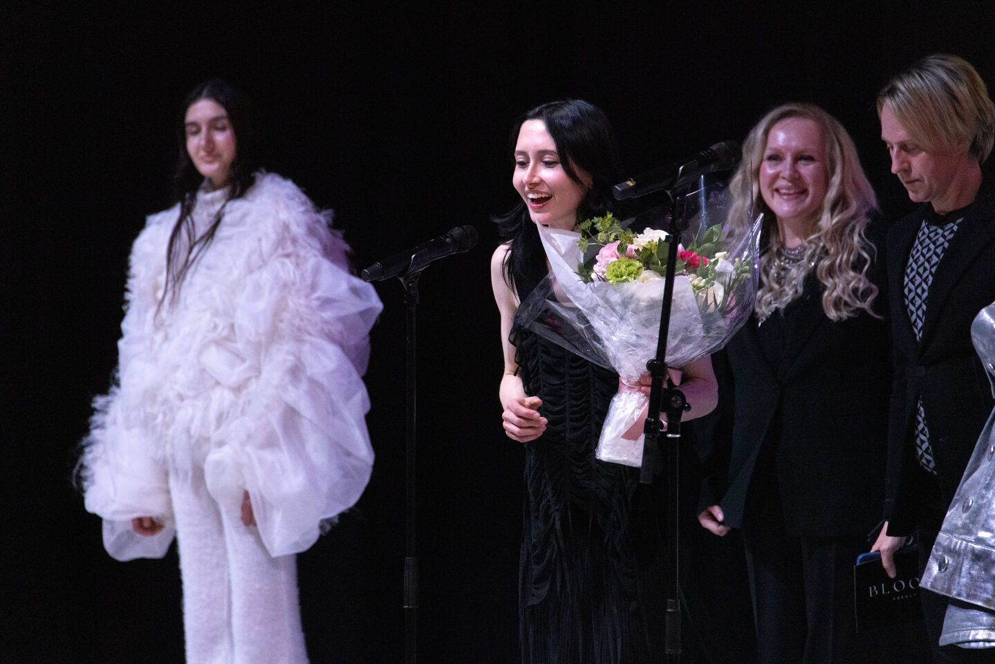 Ihmisiä lavalla; yksi valkoisessa röyhelöasussa, toinen kukkakimpun kanssa ja kaksi mustissa vaatteissa mikrofonien kanssa.