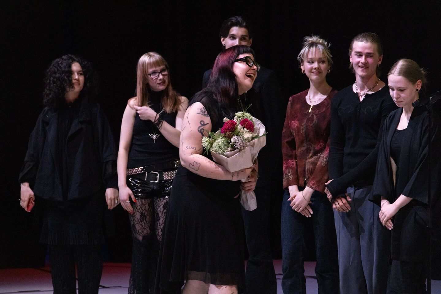 Tatuoitu nainen mustassa mekossa pitää kukkakimppua, ympärillään tummiin pukeutuneita ihmisiä lavalla.