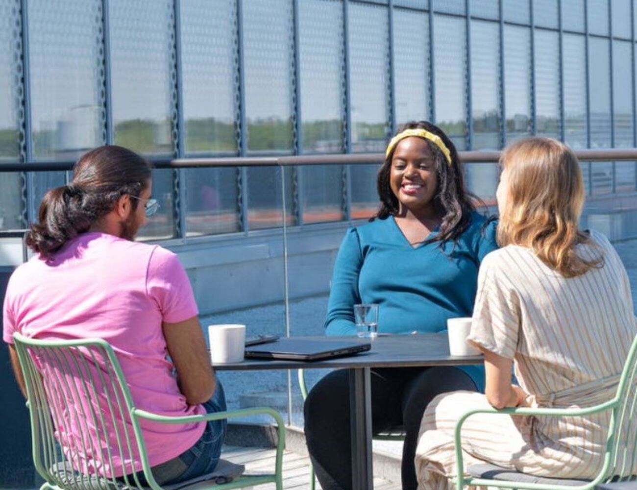 Three students having a meeting outside, sitting at a table with mugs. A laptop is on the table.
