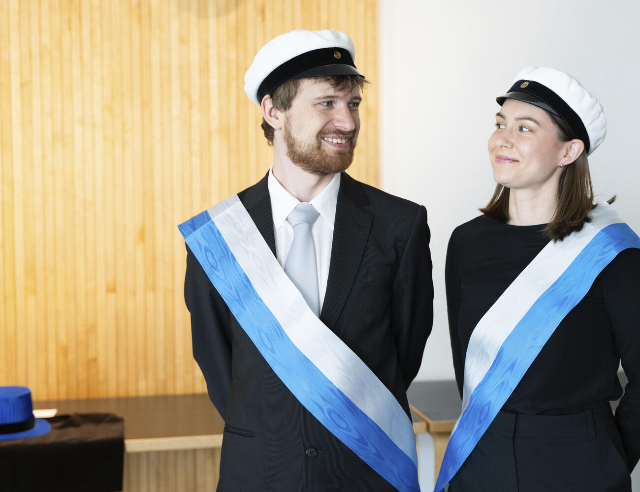 Two people wearing academic caps and black formal attire with white and blue sashes stand indoors.