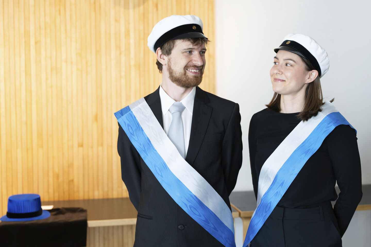 Two people wearing academic caps and black formal attire with white and blue sashes stand indoors.