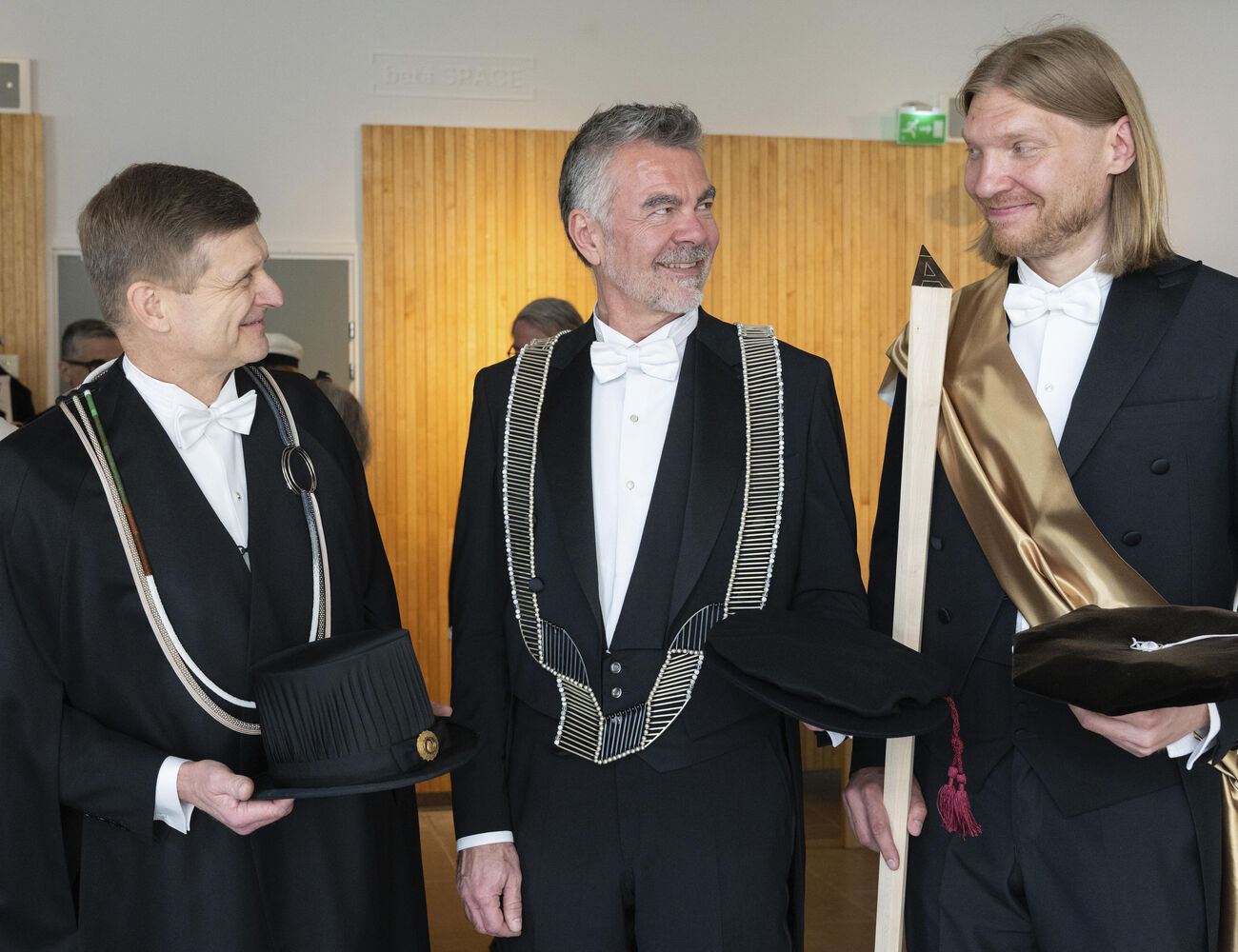 Three men in ceremonial academic garments holding formal hats and props in an indoor setting.