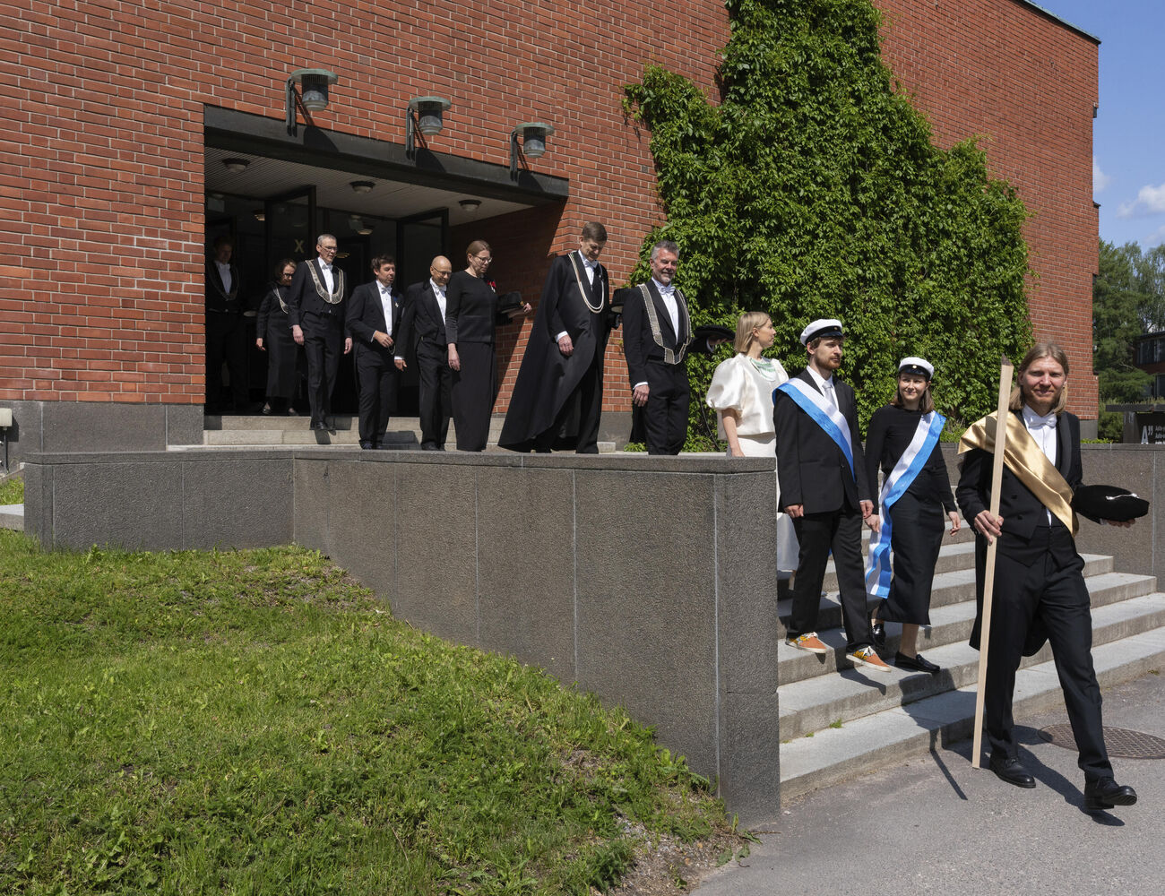 A group of people in formal attire walking down steps outside a brick building with green ivy.