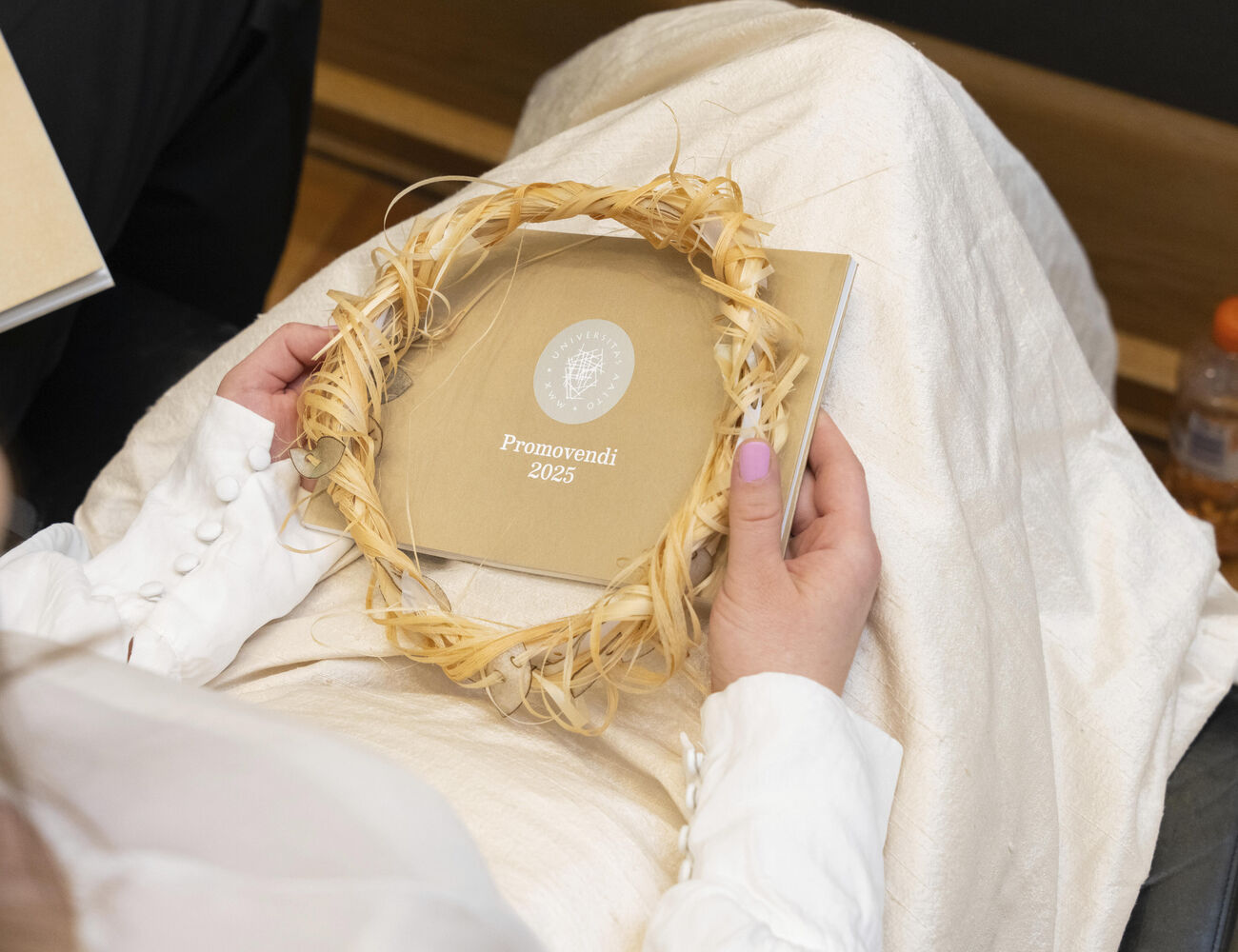 Person holding a booklet titled 'Promovendi 2023' and a straw wreath on their lap, with a bottle on the floor nearby.