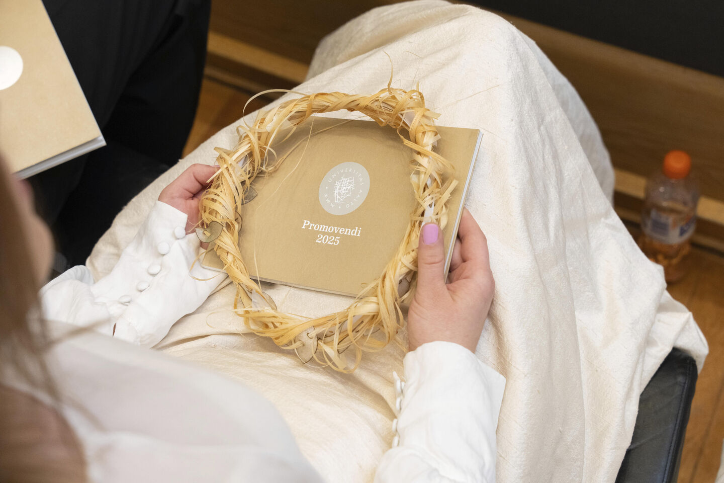 Person holding a booklet titled 'Promovendi 2023' and a straw wreath on their lap, with a bottle on the floor nearby.
