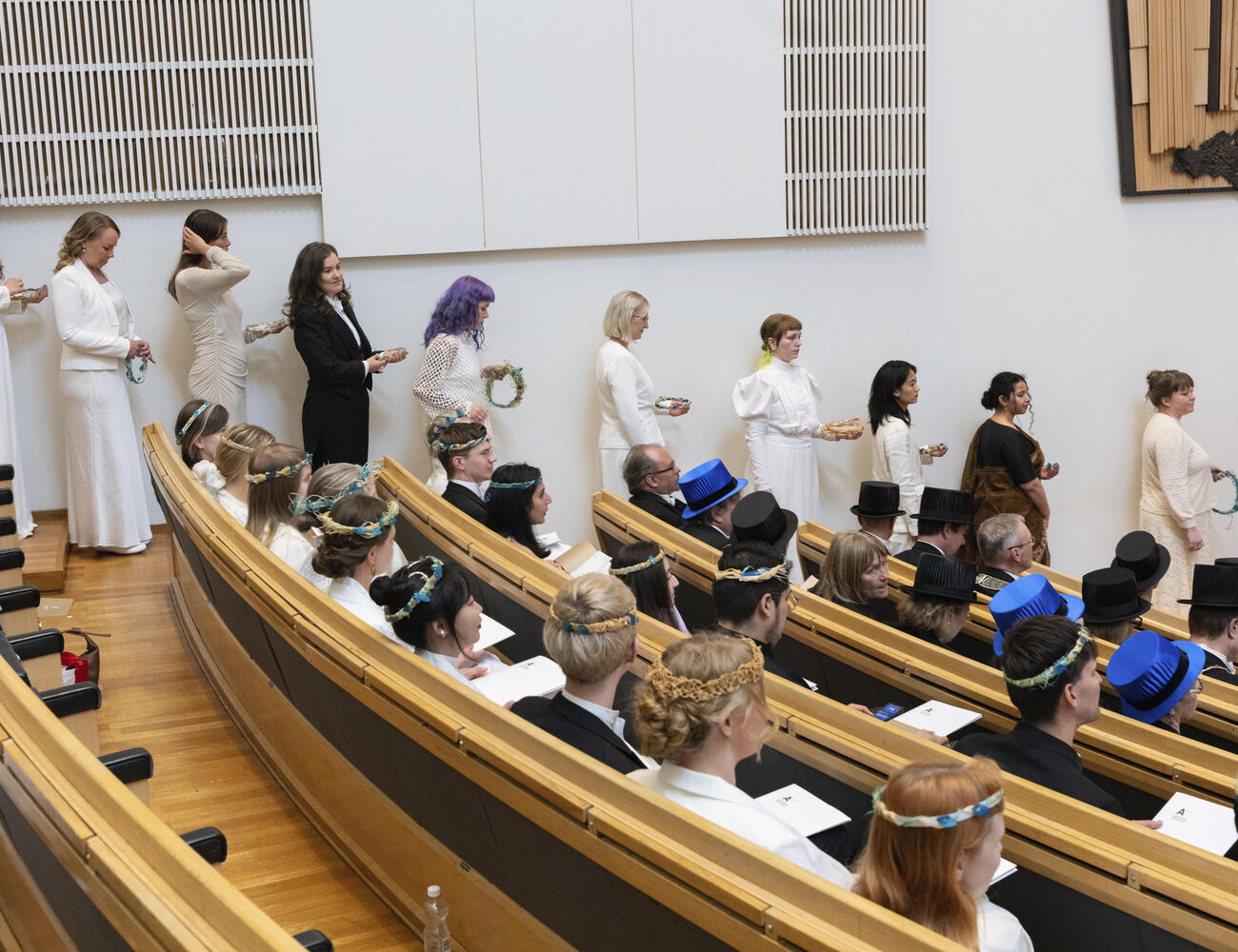 People in white academic gowns and suits line up in a lecture hall with wooden seats. Some wear laurel wreaths or academic caps.