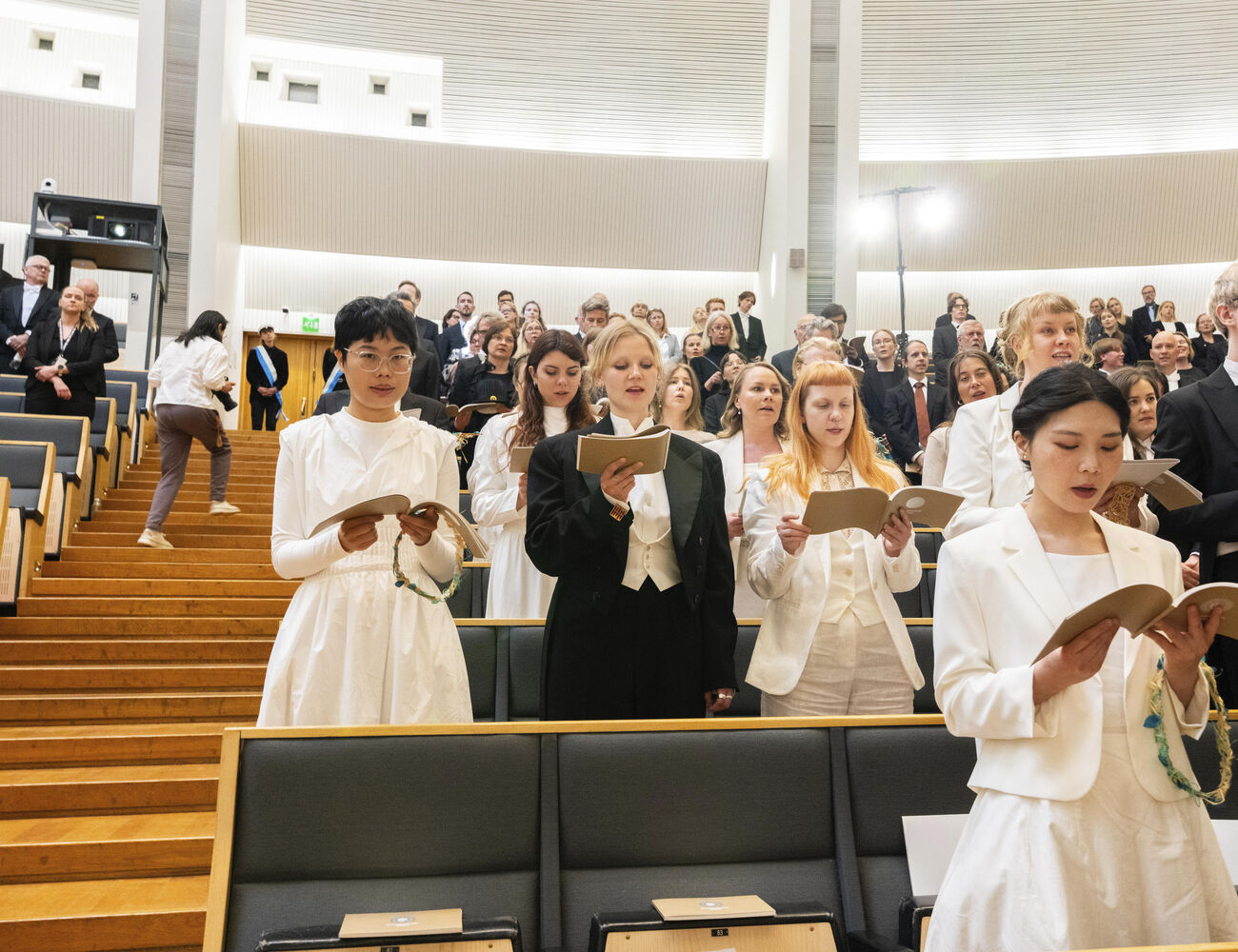 People dressed formally singing in an auditorium, holding songbooks, while others stand in the background.