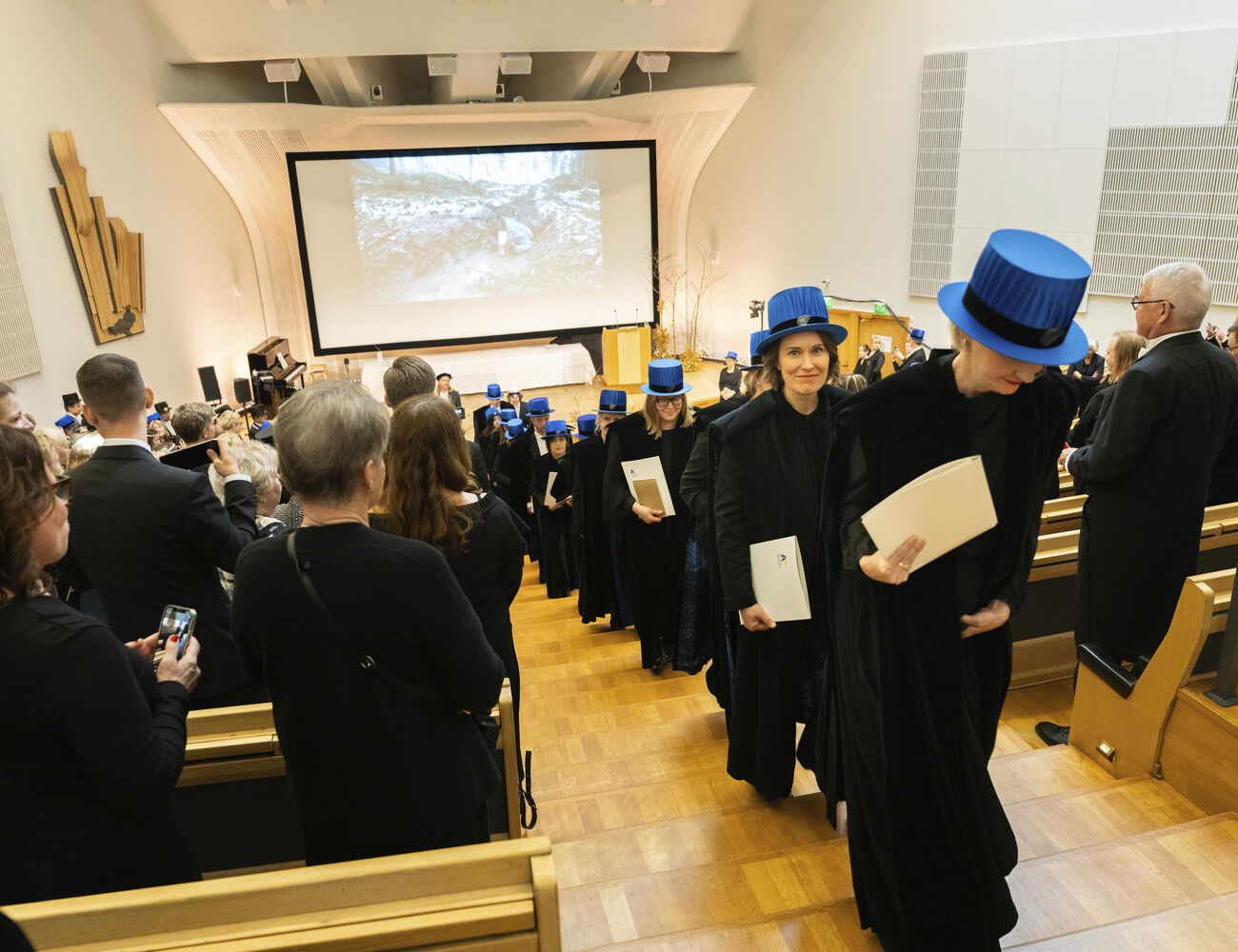 Graduation ceremony in an auditorium, graduates dressed in black gowns and blue hats holding diplomas.