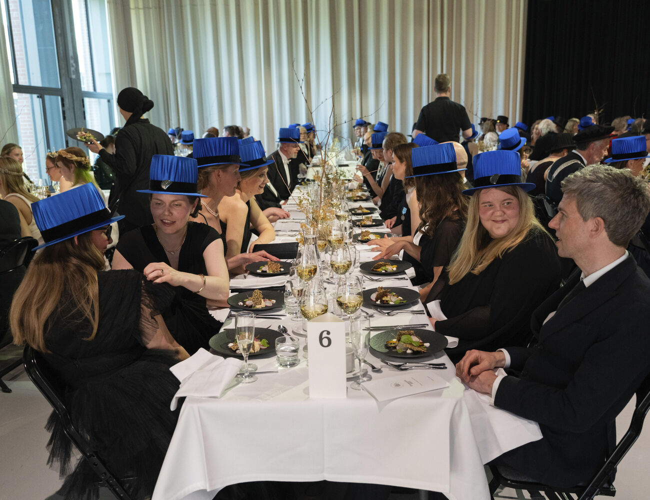 People in formal attire and blue academic hats seated at a long banquet table, number 6 card visible.