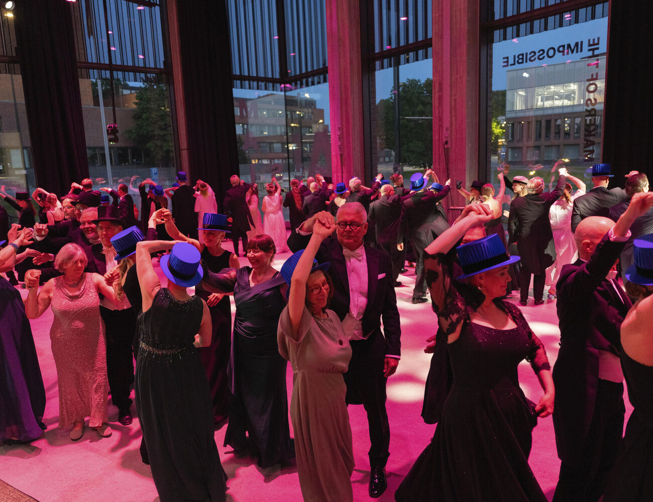 A formal event with people dancing. Many are wearing blue hats, dark evening gowns, and suits. Tall windows show an urban view.